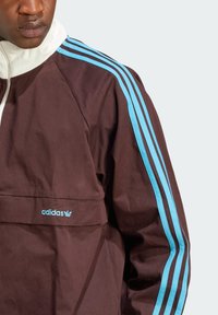 Casaco castanho com um colarinho alto, apresentando três riscas azul claras nas mangas e um logótipo bordado da Adidas na frente.