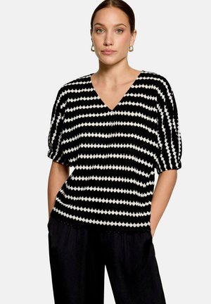 Femme portant un haut à manches courtes rayé noir et blanc avec col en V et un pantalon noir, debout les mains dans les poches devant un fond uni.