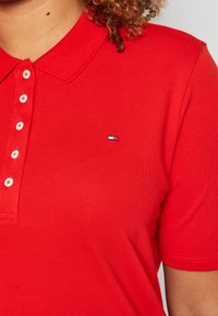 Camiseta polo roja con un tejido texturizado, cinco botones blancos y un pequeño logo bordado con acentos en azul, blanco y rojo.