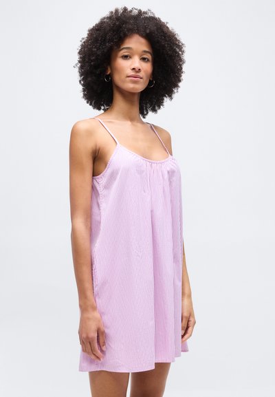 Γυναίκα με σγουρά μαλλιά που φοράει ένα φαρδύ, αμάνικο ροζ και λευκό ριγέ φόρεμα, στέκεται μπροστά σε ένα απλό φωτεινό φόντο.