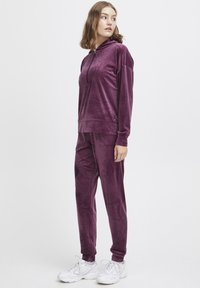 Velvet paarse trainingspak met een ritskap en een kangoeroezak en bijpassende tapered broek. Gepaard met witte sneakers.