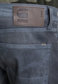 Gros plan sur la poche arrière d'un jean bleu foncé avec un patch en cuir du logo G-Star RAW et une petite étiquette métallique RAW sur le bord de la poche.