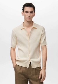 Cream short-sleeve knit shirt with a soft texture, vertical ribbed design, and a collared neckline, fastened with buttons along the front.