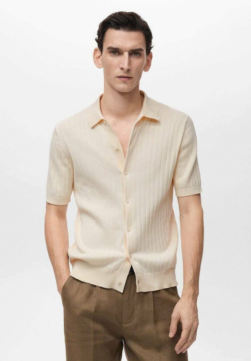 Cream short-sleeve knit shirt with a soft texture, vertical ribbed design, and a collared neckline, fastened with buttons along the front.
