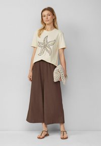 Cream-colored short-sleeve t-shirt with starfish graphic, paired with brown wide-leg pants. Holding a light crochet bag, wearing flat sandals.
