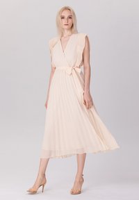 Robe enveloppante plissée pêche pâle avec des manches à volants, ceinture cintrée, et texture superposée, associée à des sandales à talons transparents.