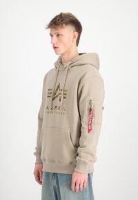 Béžová mikina s kapucňou vyrobená z mäkkej tkaniny, s logom, klokankou a vreckom na zips na rukáve s červenou visačkou.