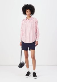 Red and white striped button-up shirt, navy shorts, black sneakers, holding a black pouch bag, standing on a white background.