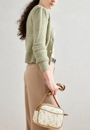 Cardigan tricotat verde deschis cu mâneci bufante, asociat cu pantaloni bej. Ținând o mică geantă cu un model floral și un bretele bej.