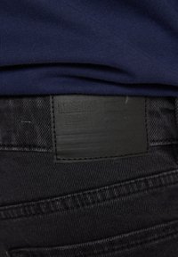 Primer plano de la cintura de unos vaqueros negros con una etiqueta de parche negro cosida y tela azul marino doblada encima.