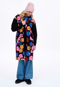 Čierny kabát s kvetinovým vzorom, ktorý má viacfarebné kvety, dlhé rukávy a zapínanie na gombík, v kombinácii s modrými širokými džínsami a čiernymi topánkami.