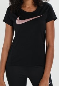 Camiseta negra de Nike, hecha de una tela ligera, con un logo metálico rosa en la parte delantera, cuello redondo y mangas cortas.