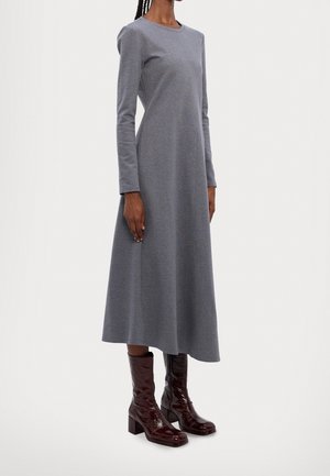 Femme portant une robe midi grise à manches longues associée à des bottes mi-mollet marron foncé et brillantes, debout devant un fond uni.
