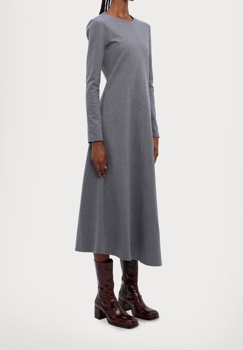 Femme portant une robe midi grise à manches longues associée à des bottes mi-mollet marron foncé et brillantes, debout devant un fond uni.