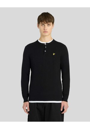 Ung mand iført en sort henley sweater med lange ærmer, tre knapper og et lille gult logo på brystet.