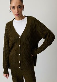 Tamsiai žalia kabelinė mezginio kardiganas su giliu V-formos iškirpimu, su šešiais dideliais šviesiais mygtukais ir plačiomis rankovėmis. Medžiaga yra tekstūruota.