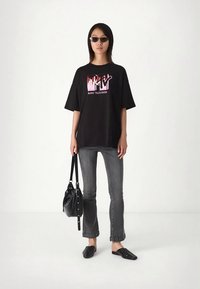 Mujer con camiseta gráfica negra de MTV, jeans grises, zapatos negros tipo slip-on, gafas de sol estilo cat-eye, sosteniendo un bolso negro, de pie en una habitación blanca.