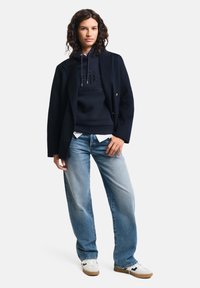 Felpa blu navy con logo in rilievo, indossata sopra una maglietta bianca, abbinata a jeans blu chiaro e sneaker bianche con dettagli marroni.