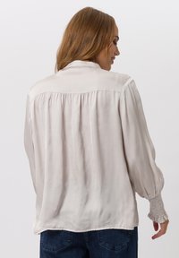 Hellbeige Bluse mit lockerem Schnitt, plissiertem Rücken und gerafften Bündchen. Aus glattem, weichem Stoff gefertigt und mit einfachem Kragen.