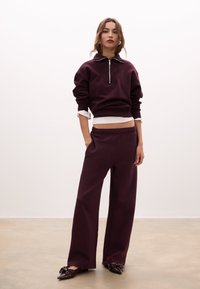 Felpa bordeaux con zip a metà, abbinata a pantaloni a gamba larga coordinati. Presenta una maglietta bianca visibile all'orlo. Tessuto morbido.