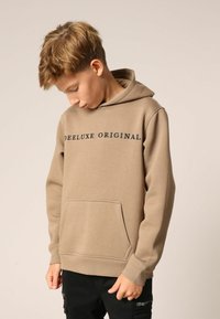 Beige hoodie met een kangoeroezak, geribde manchetten en zoom. Voorzien van "DEE LUXE ORIGINAL" in het zwart gedrukt op de borst. Zachte stoftextuur.