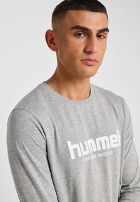 Jasnoszara, długosleeve'owa koszulka wykonana z miękkiego materiału, z białym logo "hummel" i napisem "SPORTWEAR SINCE 1923" z przodu.
