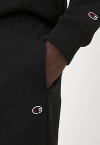 Sudadera de algodón negra con un bolsillo lateral, que presenta un pequeño logo bordado en rojo y blanco en el puño de la manga. La textura es suave y mate.