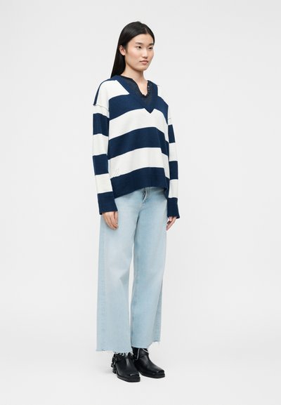 Γυναίκα με μαύρα μακριά μαλλιά φοράει μπλε-σκούρη και λευκή ριγέ πουλόβερ με V-λαιμόκοψη, ανοιχτό μπλε τζιν με φαρδύ πόδι και μαύρα μποτάκια μέχρι τον αστράγαλο, στέκεται πλευρικά.
