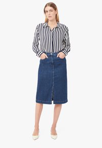 Camicia a righe bianco e nera, abbinata a una gonna di jeans blu scuro. La gonna presenta uno spacco frontale e una vita standard. Modella con tacchi beige.