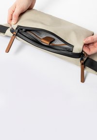 Beige Reißverschlussbeutel mit einem Lederziehband und schwarzem Netzinnenfutter. Die Hand ist so positioniert, dass der Reißverschluss geöffnet wird.
