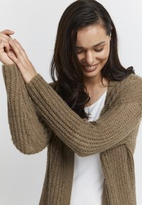 Brūns ribots garš kardigans, izgatavots no struktūrizētas dzijas. Atvērta priekšējā daļa ar nolaižamām plecu daļām un platām piedurknēm, uzvilkts virs baltas blūzes.