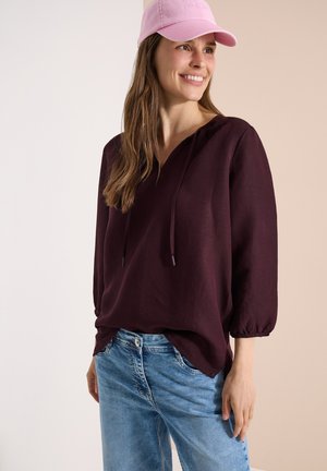 Uśmiechnięta kobieta, ubrana w różową czapkę baseballową, luźną ciemnofioletową bluzkę z rękawami do połowy przedramienia oraz jasnoniebieskie dżinsy na neutralnym tle.