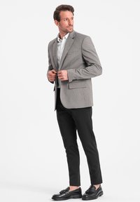 Grau karierter Blazer mit Reverskragen, zwei Fronttaschen und schwarzen Anzughosen. Schwarze Slipper vervollständigen das Outfit. Darunter ein weißes Hemd.