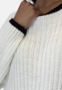 Hvid strikket sweater med ribbet struktur, med sort krave og manchetter. Nærbillede, der viser detaljer af materialet og syningerne.
