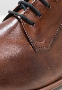 Chaussure en cuir marron avec une finition lisse, dotée de lacets noirs, d'un bout pointu et de surpiqûres détaillées sur les côtés.