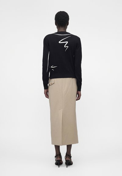 Γυναίκα που φοράει μαύρο πουλόβερ με λευκές αφηρημένες γραμμές, μπεζ midi φούστα με πλαϊνή τσέπη και μαύρα ψηλοτάκουνα σανδάλια, στέκεται με την πλάτη γυρισμένη.