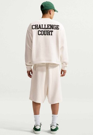 Mężczyzna w białej koszuli z długim rękawem z tekstem "CHALLENGE COURT", białych szortach, zielonej czapce, białych skarpetkach i zielonych butach sportowych, odwrócony tyłem.