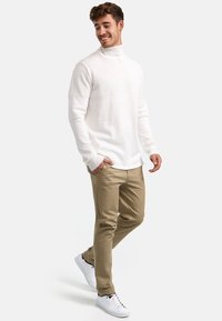Hvit, teksturert genser med høy hals som er kombinert med beige bukser og hvite sneaker. Genseren har en ribbestrikket krage og subtile horisontale mønstre.