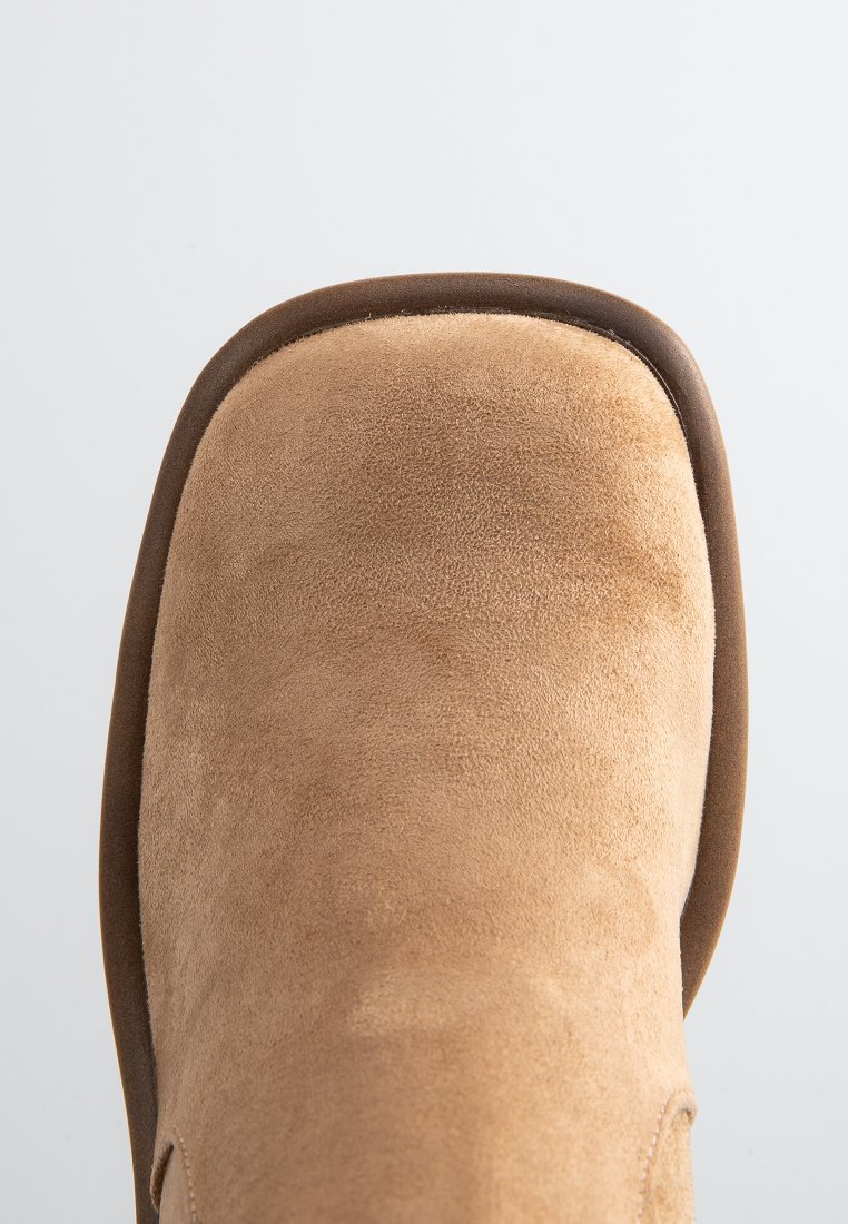 Bottine en suédine beige avec un bout arrondi et une bordure marron. La texture est douce, avec une finition lisse sans motifs ni ornements.