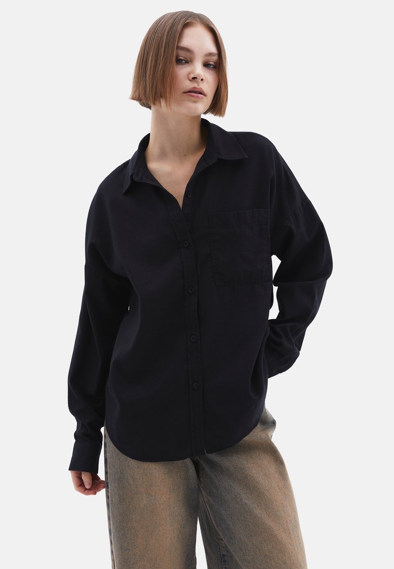 Zwarte knoopshirt met een ontspannen pasvorm, kraag, enkele borstzak en lange mouwen, gecombineerd met beige brede broek.
