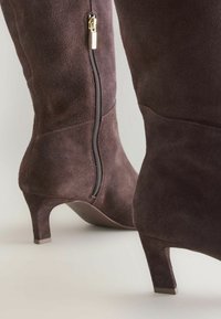 Brown suede ankle boots with a mid-height heel, featuring a side zipper and a smooth texture, emphasizing a sleek, fitted design.