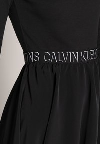 Black dress featuring a ribbed bodice and flowing skirt. The waistband displays "CALVIN KLEIN" in white embroidery. Smooth, textured fabric.
