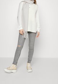 Zapatillas altas beige con suelas blancas, jeans skinny grises con detalle desgastado, camisa blanca de botones y bufanda de punto gris claro.
