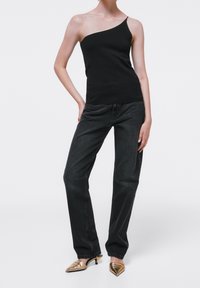 Top nero a spalla unica abbinato a jeans di denim scuri; la modella indossa mules dorate a punta. Texture liscia, vestibilità rilassata, design minimal.