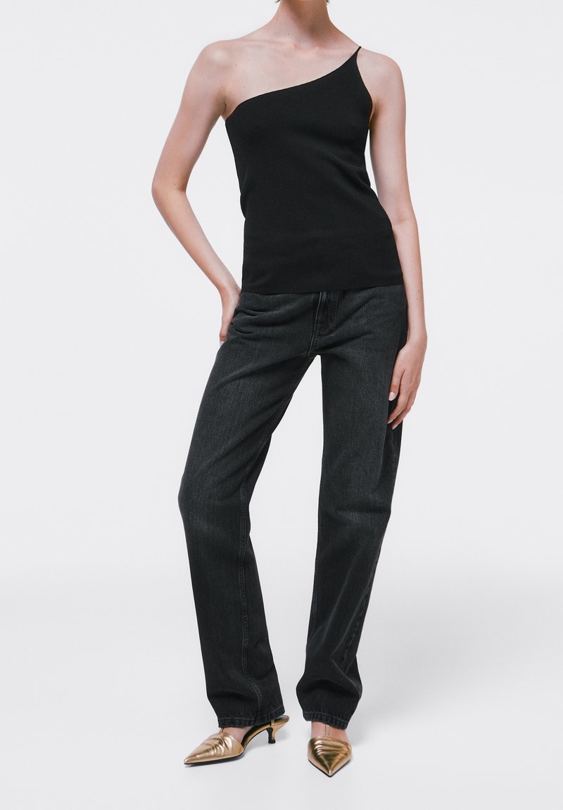 Top nero a spalla unica abbinato a jeans di denim scuri; la modella indossa mules dorate a punta. Texture liscia, vestibilità rilassata, design minimal.