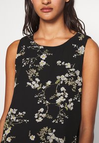 Robe sans manches noire ornée de broderies florales blanches et jaunes avec des accents verts. Encolure ronde et tissu doux et texturé.