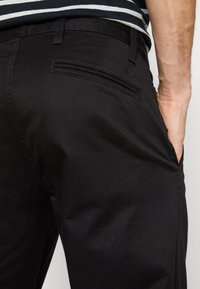 Homme portant un pantalon noir avec une poche passepoilée à l'arrière et la main dans la poche latérale, associé à une chemise rayée noire et blanche.