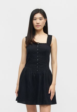 Γυναίκα με ίσια σκούρα μαλλιά, φορώντας αμάνικο μαύρο φόρεμα με κουμπιά στο μπροστινό μέρος, στέκεται μπροστά σε ένα απλό φωτεινό φόντο.