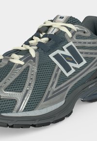 Σκούρο γκρι αθλητικό παπούτσι New Balance με λευκά κορδόνια, επάνω μέρος από δίχτυ και παχιά λαστιχένια σόλα με λογότυπο της μάρκας στο πλάι.