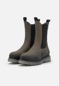Botas de tobillo de goma en verde oscuro con paneles elásticos negros, talón acolchado y suela gruesa y transparente. Textura lisa con costuras mínimas.